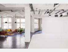 Großzügiger, heller Loft-Raum mit einer Sitzecke, bestehend aus Holzmöbeln, bunten Kissen und Zimmerpflanzen, neben einem Fotostudio-Bereich mit weißem Hintergrund und professionellen Beleuchtungsanlagen an der Decke. Große Fenster lassen viel Tageslicht einfallen, die Wände und Decken sind weiß gestrichen.