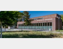 Backsteingebäude der Rettungswache Ludwigsfelde mit großen Glasflächen an der Vorderseite, umgeben von Grünflächen und Bäumen. Im Vordergrund eine gepflasterte Fläche und vereinzelte Straßenlaternen.