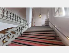 Eine breite Treppe mit roten Stufen und weißen Stufenrändern in einem historischen Gebäude, flankiert von einem geschwungenen, verzierten Geländer links und einfachen Metallhandläufen. Im Hintergrund sind hohe Rundbögen, ein Kronleuchter und eine Reihe von Fenstern zu sehen.
