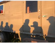 Schatten von fünf Personen sind auf der sonnenbeschienenen Außenwand eines hellbraunen, fenster- und türlosen Anhängers zu sehen. Im Vordergrund befinden sich eine Reifenabdeckung und ein Holzblock, während die Umgebung grasbedeckt ist.