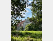 Historisches Gebäude mit barockem Giebel und roten Ziegeldächern, umgeben von dichtem Grün und Bäumen im Vordergrund; teilweise blauer Himmel mit Wolken im Hintergrund.