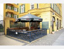 Außenbereich eines Restaurants mit gelber Fassade und grünen Fensterläden. Die Terrasse ist durch ein blaues Metallgeländer abgetrennt und mit Holzbänken und -tischen sowie einem großen Sonnenschirm mit der Aufschrift "Berliner Pilsner" ausgestattet.