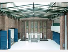 Großer, moderner Innenraum mit Backsteinwänden, großen vergitterten Fenstern und einem Glasdach. Der Boden ist weiß, Türen und Elemente an den Wänden sind blau, und in der Mitte befindet sich eine Glasdrehtür.