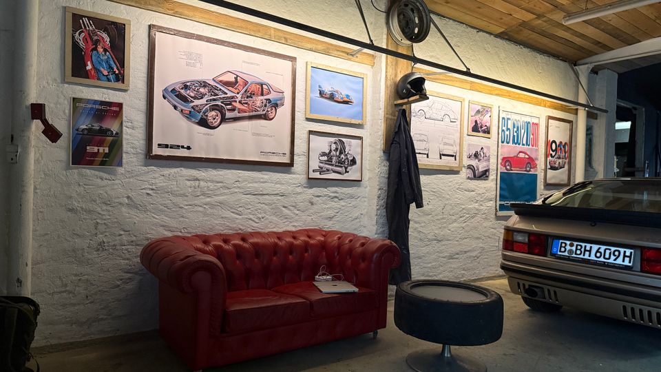 Innenraum einer Garage mit weiß verputzten Wänden und Holzbalkendecke, dekoriert mit gerahmten Auto-Postern und technischen Zeichnungen. Vor der Wand steht ein rotes Ledersofa mit kleinem Beistelltisch, daneben ist das Heck eines geparkten Autos zu sehen.