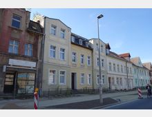 Straßenszene mit einer Reihe von älteren Wohnhäusern in unterschiedlichen Pastellfarben, teils renovierungsbedürftig. Im Vordergrund sind Baustellenabsperrungen und ein unvollendeter Gehweg sichtbar, daneben eine Person auf einem Fahrrad und eine Straßenlaterne.