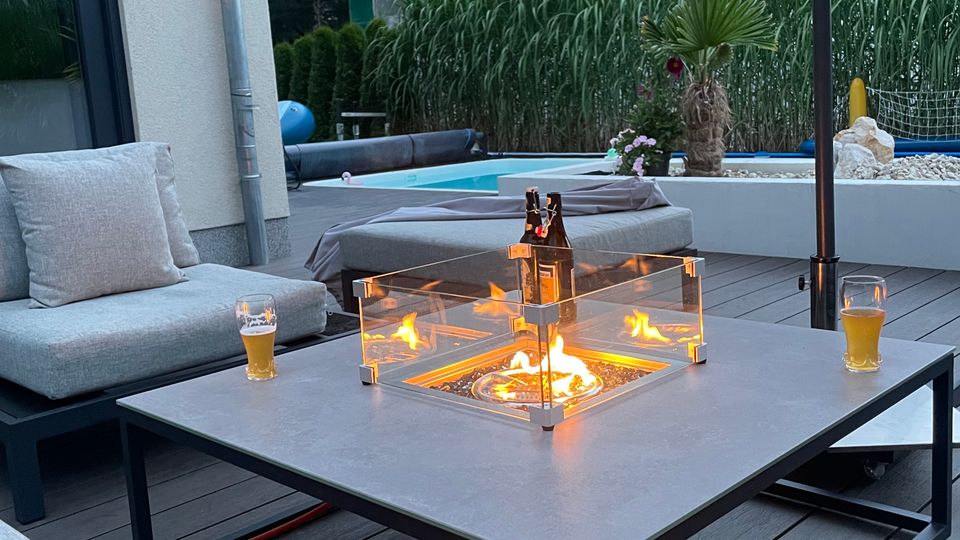 Moderne Terrasse mit grauer Holzoptik, einer Feuerstelle in einem rechteckigen Tisch mit Glasumrandung und Sitzgelegenheiten mit hellgrauen Polstern. Im Hintergrund sind hohe Ziergräser, ein Pool sowie ein mit Baugerüsten verkleidetes Gebäude zu sehen.