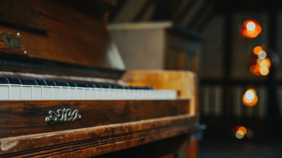 Nahaufnahme eines hölzernen Klaviers mit sichtbarer Tastatur und verziertem Metallschild an der Vorderseite. Im Hintergrund ein unscharfer Innenraum mit warmen, runden Lichtquellen und angedeuteten Deckenbalken.