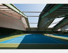 Ein überdachtes Tenniscenter mit blau-grünen Tennisplätzen, Metallträgern und teilweise offenen Dachelementen, durch die Tageslicht einfällt. Die Halle ist großzügig gestaltet und enthält Beleuchtung an den Deckenträgern.