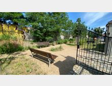 Kleiner Parkbereich mit Sandboden, Holzbänken und vereinzelter Vegetation, eingefasst von einer Betonmauer mit Graffiti. Im Vordergrund ein geöffnetes schmiedeeisernes Tor, im Hintergrund dichte Bäume und Büsche.