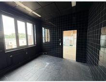 Leerer Innenraum mit schwarz gefliesten Wänden und Boden, abgehängter Decke mit Leuchtstoffröhre und großem Fenster zur Außenansicht. Rechts führt eine Türöffnung in einen helleren, ebenfalls gefliesten Nebenraum.