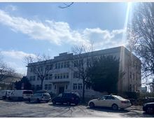 Ein dreistöckiges, rechteckiges Gebäude mit weißer Fassade und großen Fenstern im Stil des 20. Jahrhunderts, umgeben von kahlen Bäumen. Im Vordergrund stehen parkende Autos entlang einer Straße, der Himmel ist leicht bewölkt mit Sonnenschein.