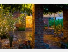 Gemütlicher Innenhof mit Backsteinboden und -wänden, umgeben von blühenden Pflanzen und Büschen. Im Vordergrund sind ein Holzsitzbereich mit Sonnenschirm und eine Laterne zu sehen, während durch die Fenster eines Gebäudes warmes Licht scheint.