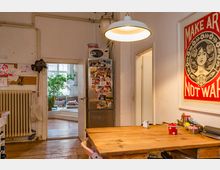 Wohnküche mit Holzboden und rustikalem Holztisch, im Hintergrund ein Raum mit Sofa und Pflanzen. An der Wand ein rotes Kunstposter, an der Kühlschranktür verschiedene Fotos und Magnete angebracht.