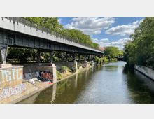 Eine stählerne Brücke erstreckt sich über einen mit Graffiti dekorierten Uferbereich entlang eines begradigten Flusses, umgeben von Bäumen und Grünflächen. Helle Wolken und blauer Himmel verleihen der Szene eine offene Atmosphäre.