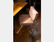 Ein moderner Drehstuhl mit beigefarbenem Stoffbezug und Metallrahmen steht neben einem Holztisch. Der Boden besteht aus Parkett, auf dem teils ein beigefarbenes Fell liegt.