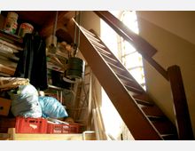Ein Abstellraum mit einer steilen Holztreppe, die zu einem oberen Bereich führt, daneben ein bunt verglastes Fenster, das Licht hereinlässt. Der Raum ist mit Regalen, gefüllten Kisten und Gegenständen wie Kleidersäcken und Leitern unordentlich eingerichtet.