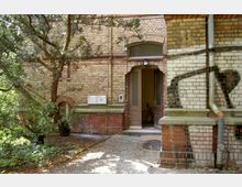 Backsteinfassade eines Gebäudes mit rundbogigen Fenstern und einer geöffneten Holztür, die in einen Innenraum mit einem Fahrrad führt. Im Vordergrund befindet sich ein kleiner gepflasterter Weg und ein Baum, rechts ist ein Regenrohr an der Wand zu sehen.