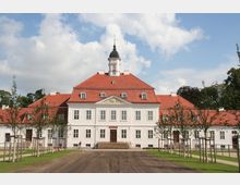 Barockes Herrenhaus mit weißer Fassade und rotem Ziegeldach, zentral gekrönt von einem Uhrenturm mit Kuppel. Der Vordergrund zeigt eine breite Zufahrt und junge Bäume, die entlang eines Gehwegs gepflanzt sind.