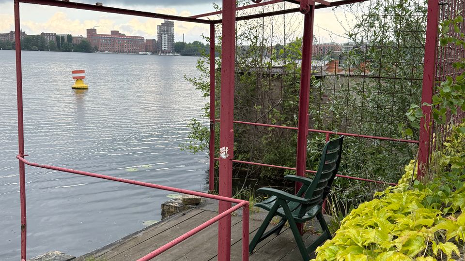 Überwucherter Uferplatz an einem See mit einem roten Metallgerüst über einem kleinen Holzdeck und Geländer. Im Hintergrund Wasserfläche mit Boje und gegenüberliegender Uferbebauung unter bewölktem Himmel.