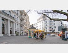 Straßenszene mit einem kleinen gelben Kiosk in der Mitte, umgeben von Souvenirständen; im Hintergrund historische Gebäude mit verzierten Fassaden und moderne weiße Gebäude. Ein Baum mit kahlen Ästen ragt ins Bild, und im rechten Bildbereich stehen Straßenlampen und Fahrräder.