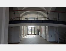 Heller Durchgangsbereich mit gewölbter Decke und einer modernen Brücke mit Glasgeländer, die den oberen Bereich verbindet. Im Hintergrund eine große Glastür, die zu einem Außenbereich mit Rasenflächen führt.