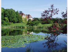 Großer Teich mit Seerosen im Vordergrund, umgeben von dichter Vegetation und einem Baum mit roten Blättern rechts. Im Hintergrund ein mehrstöckiges Backsteingebäude mit roten Dächern, eingebettet in eine grüne Wiesenlandschaft.