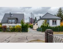 Zwei Einfamilienhäuser mit Satteldächern und weißer Fassade, umgeben von Zäunen und gepflegten Gärten; im Hintergrund sind Industriekamine zu sehen. Der Himmel ist bewölkt, und die Szenerie zeigt eine ruhige Wohngegend.