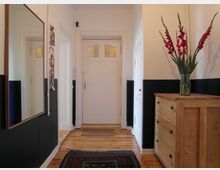 Flur mit hell gestrichenen Wänden, deren unterer Bereich schwarz abgesetzt ist, und einer Holztür mit Glaseinsätzen. Der Raum enthält einen großen Spiegel an der Wand, eine Holzkommode mit einer Vase und roten Blumen sowie einen Teppich auf einem Holzboden.