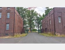 Zwei backsteinerne Gebäude mit symmetrischem Design stehen auf beiden Seiten einer asphaltierten Einfahrt. Im Hintergrund wachsen große Bäume, und ein Metalltor begrenzt den Zugang.