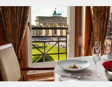 Blick aus einem eleganten Restaurant durch ein Fenster mit dekorativen Vorhängen auf das Brandenburger Tor in Berlin. Im Vordergrund ein gedeckter Tisch mit einem Teller Essen, Weingläsern und einer Schale mit roten Rosen.