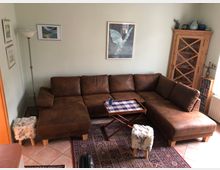 Wohnzimmer mit einer braunen Ledercouch in L-Form, einem kleinen Holztisch mit blau-weiß karierter Tischdecke und zwei Hockern mit Fellbezug. Im Hintergrund befinden sich eine Holzvitrine, eine Stehlampe, gerahmte Bilder an der Wand und ein Teppich auf Fliesenboden.