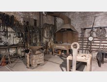 Historische Schmiede mit einem Amboss auf einem Holzsockel, einem Schmiedeofen mit Abzugshaube, sowie Wänden voller aufgehängter Werkzeuge und Metallketten. Im Raum befinden sich außerdem ein großer Schraubenschlüssel auf einem Holztisch und eine Rundskala an der Wand.