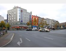 Straßenszene mit einem breiten Kreuzungsbereich, umgeben von mehrstöckigen Gebäuden mit Geschäften im Erdgeschoss, darunter ein Hotel und eine Apotheke. Im Vordergrund sind mehrere Fahrzeuge, darunter Taxis, sowie auf der rechten Seite ein U-Bahn-Eingang erkennbar.