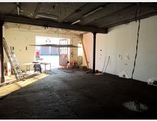 Ein leerer, industrieller Innenraum mit rauem Betonboden, freiliegender Decke und großen offenen Doppeltüren, die Tageslicht hereinlassen. An der weißen Wand hängen Zeichnungen, und im Raum befinden sich Arbeitsmaterialien, ein Tisch, eine Leiter sowie vereinzelte Werkzeuge.