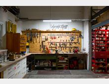 Eine Werkstatt mit einer beleuchteten Arbeitsfläche, an der Wand hängen verschiedene Handwerkzeuge wie Sägen, Schraubenzieher und Zangen ordentlich sortiert. Rechts stehen rote Regale mit zahlreichen Kleinteilen, während die Arbeitsfläche unten mit Schubladen und Regalen ausgestattet ist, die weitere Werkstattmaterialien und Zubehör enthalten.