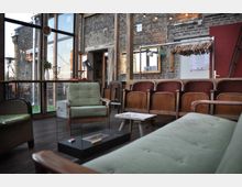 Ein gemütlicher Raum mit einer Mischung aus Holz- und Backsteinwänden, großen Fenstern, die nach außen blicken, und einer Einrichtung aus Polstermöbeln und alten Holztheaterstühlen. Im Vordergrund stehen grüne Sessel, ein kleiner Holzhocker und ein moderner Tisch, während an einer Wand Kleiderbügel sowie eine Dekoration mit Lichterketten zu sehen sind.