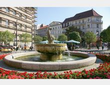 Brunnen auf einem gepflasterten Platz, umgeben von Blumenbeeten mit roten Blumen und schattenspendenden Bäumen; im Hintergrund befinden sich mehrstöckige Gebäude, darunter eines mit Baugerüst, sowie Sitzbereiche mit Sonnenschirmen.