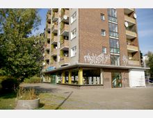 Mehrstöckiges Wohngebäude mit Klinkerfassade und Balkonen, im Erdgeschoss befindet sich eine Bücherei mit großen Schaufenstern. Im Vordergrund sind ein gepflasterter Platz, Pflanzenkübel und ein Baum sichtbar. Graffiti bedecken Teile der Fassade.