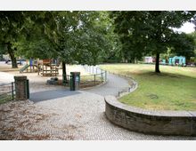 Spielplatz und Parkanlage mit Bäumen, einer geschwungenen gepflasterten Gehwegfläche, einer Kletterstruktur aus Holz mit Rutsche und einer angrenzenden Wiese; im Hintergrund ein blaues Pavillon und Gebäude.