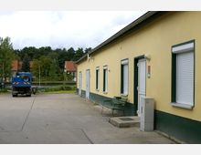 Gelbes Gebäude mit grün umrahmten Fenstern, geschlossenen Rollläden und mehreren Türen, daneben eine grüne Bank. Im Hintergrund sind rote Ziegeldächer, Bäume und ein Wasserlauf zu sehen. Links steht ein blauer Lastwagen auf einem asphaltierten Hof.