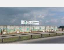 Eine Betonwand mit bemalten Bildern von Bäumen und Landschaften steht entlang einer Straße, geschützt durch eine Leitplanke. Darüber befindet sich ein großes Schild mit der Aufschrift „Tiefbau- und Schüttguthandel“ und Kontaktdaten.