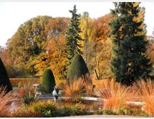 Ein gepflegter Garten mit herbstlicher Bepflanzung, darunter geschnittene Buchsbaumkegel, Ziergräser und ein kleiner Teich mit einer modernen skulpturalen Dekoration in der Mitte. Im Hintergrund stehen Bäume mit buntem Herbstlaub und immergrüne Nadelbäume.