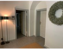 Wohnungsflur mit hellen Wänden und einem gefliesten Boden. Zu sehen sind eine geöffnete weiße Tür, ein beleuchteter Stehlampen-Doppelkopf, ein Intercom-Gerät an der Wand und ein rundes Dekokranz-Element.