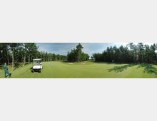 Panoramaansicht eines Golfplatzes, umgeben von dichtem Wald. Im Vordergrund eine gepflegte Rasenfläche, ein weißes Golfcart und eine Person in Arbeitskleidung auf der linken Seite.