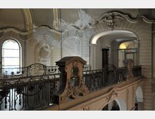 Barocker Innenraum mit einer verzierten Balustrade aus Schmiedeeisen, darüber geschwungene Stuckelemente und ein Relief eines Adlers. Im Hintergrund sind hohe Fenster mit ornamentalem Glas sowie Rundbögen in heller Wandgestaltung sichtbar.