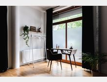 Minimalistisch eingerichteter Raum mit Holzboden, einem Schreibtisch mit Stuhl vor großen Fenstern mit halbtransparentem Sichtschutz und schwarzen Vorhängen. An der linken Wand befindet sich ein Kleiderständer, ein Heizkörper sowie ein Wandregal mit Pflanzen und Dekorationen; eine Zimmerpflanze steht rechts im Vordergrund.