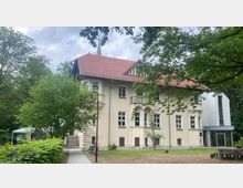 Zweistöckiges Gebäude mit beiger Fassade und rotem Satteldach, umgeben von Bäumen und Grünflächen. Ein moderner weißer Gebäudeteil mit Glasfront befindet sich rechts, während ein gepflasterter Weg und eine Sitzbank im Vordergrund sichtbar sind.