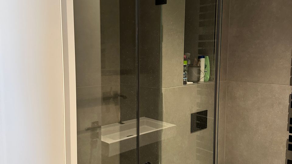 Modernes Badezimmer mit grauen Fliesen und einer bodengleichen Glasdusche mit schwarzem Rahmen und Regendusche. Rechts ist ein wandhängendes WC zu sehen sowie eine eingelassene Wandnische für Pflegeprodukte.