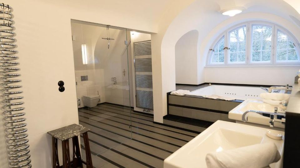 Modernes Badezimmer mit gewölbter Decke, gestreiftem Fliesenboden und großem Rundbogenfenster. Rechts stehen eine eingelassene Eckbadewanne und ein Doppelwaschbecken; links befindet sich eine verglaste Dusche mit Handtuchheizkörper.