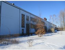 Außenansicht eines schlichten, länglichen Industriegebäudes mit weißer Fassade und rechteckigen Fenstern. Im Vordergrund liegt eine schneebedeckte Fläche mit einzelnen kahlen Bäumen und Pflanzen, während der Himmel klar und blau ist.
